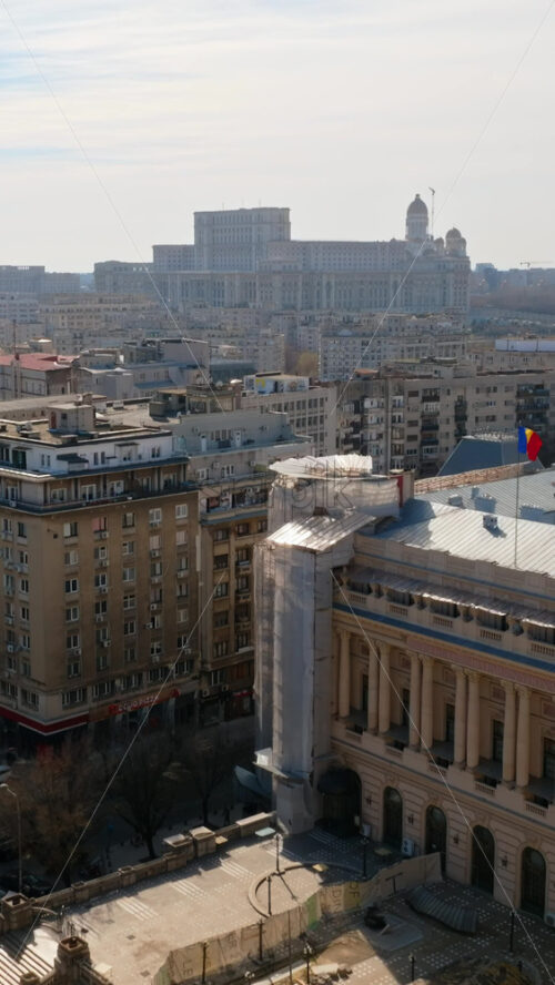 Video - Bucharest, Romania - March 03, 2024: Vertical aerial drone view of a downtown road with moving traffic and walking people. Palace of the Parliament in background. Sunny day