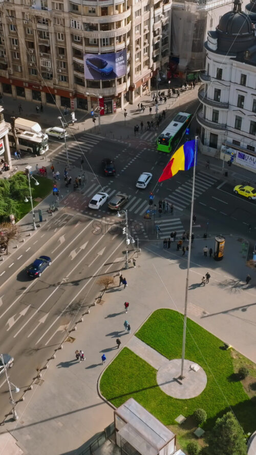 Video - Bucharest, Romania - March 03, 2024: Vertical aerial drone view of a downtown intersection with moving traffic and walking people in sunlight. Waving national flag