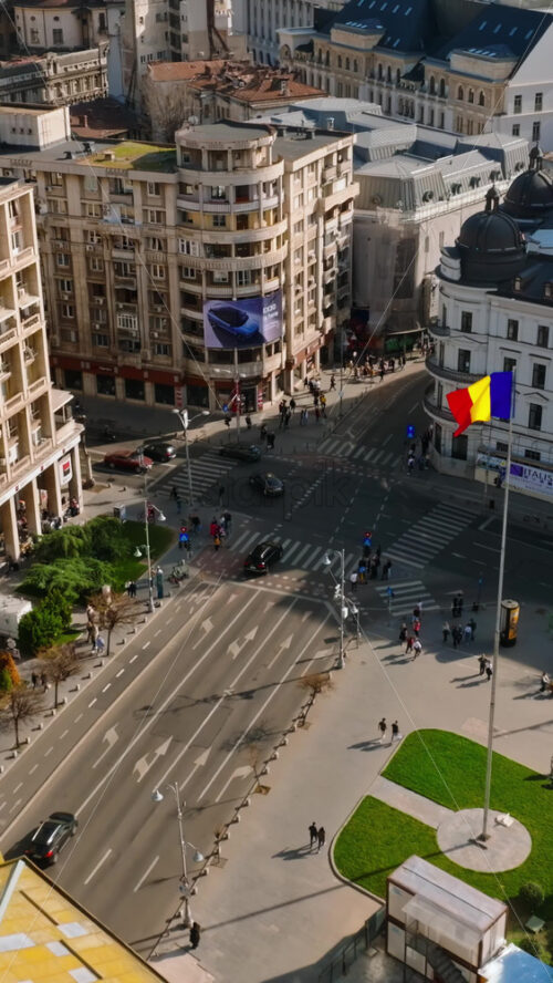 Video - Bucharest, Romania - March 03, 2024: Vertical aerial drone view of a the city center with historical buildings, moving traffic and walking people in sunlight. Waving national flag