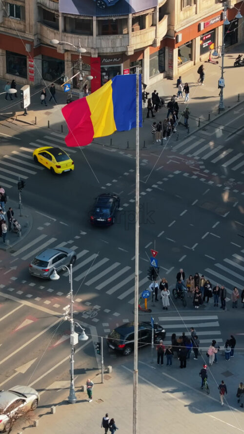 Video - Bucharest, Romania - March 03, 2024: Vertical aerial drone view of a downtown intersection with moving traffic and walking people in sunlight. Waving national flag