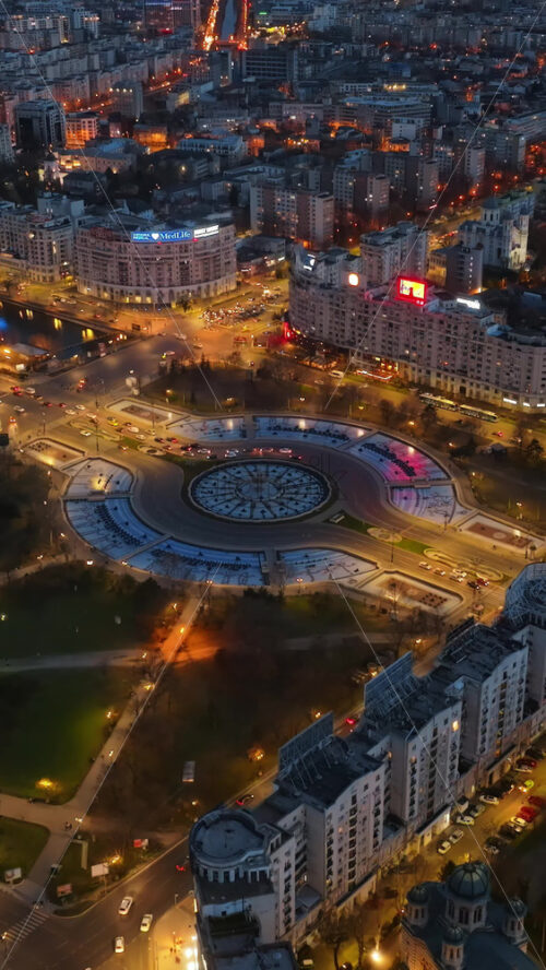 Video - Bucharest, Romania - March 03, 2024: Vertical aerial drone view of the illuminated city downtown in the evening. Roundabout intersection with moving cars and fountains. Blue hour