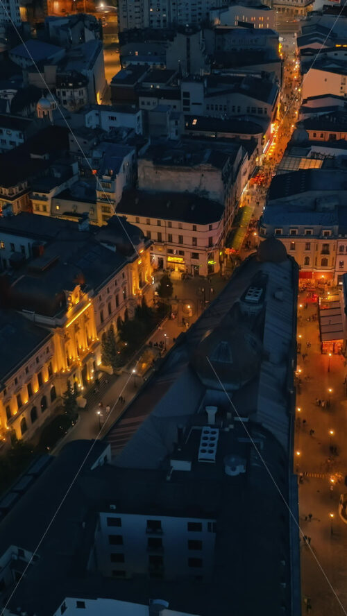 Video - Vertical aerial drone view of walking people among old historical buildings and commercial shops in the old town in the evening. Blue hour in Bucharest, Romania