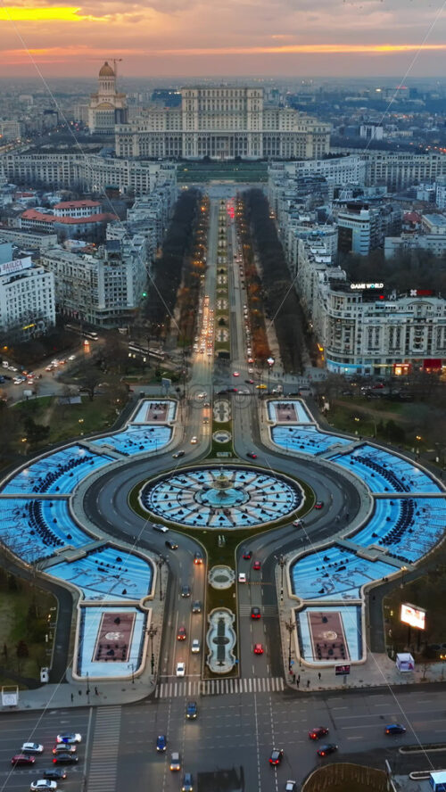 Video - Bucharest, Romania - March 03, 2024: Vertical aerial drone view of the city downtown at sunset. Roundabout intersection with moving cars and fountains. Palace of the Parliament in the distance