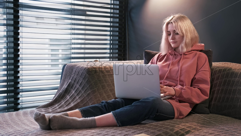 Video - Young content creator blonde girl is on her laptop sitting on the sofa near the window. Working from home