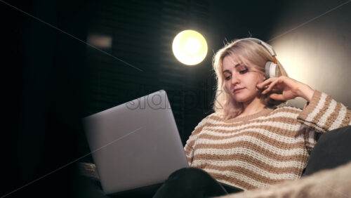 Video - Young content creator blonde girl with headphones working on her laptop sitting on the sofa