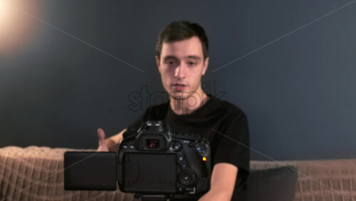 Video - Young content creator talking and gesturing man filming himself using a camera on a tripod while recording a vlog. Working from home