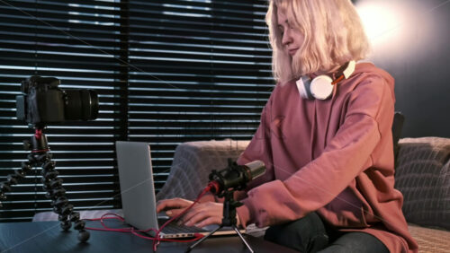 Video - Young content creator blonde girl with headphones working on her laptop on the table with camera on a tripod, microphone. Working from home