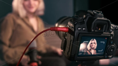 Video - Young content creator blonde girl connecting a wire to the camera on a tripod and filming herself talking for vlog. Working from home
