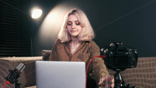 Video - Young content creator blonde girl with cup of water sitting down on the sofa, starting working on her laptop, camera on a tripod, microphone. Working from home