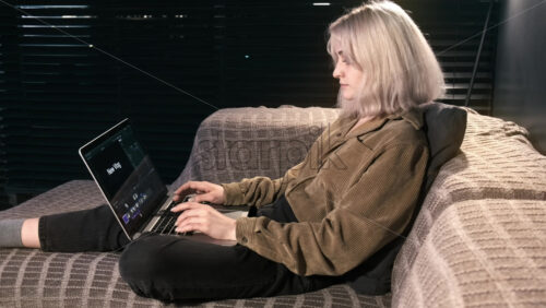Video - Young content creator blonde girl is on her laptop sitting on the sofa. Working with video from home