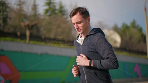 Video - Young focused man running in jacket. Moving cars. Cloudy weather. Slow motion