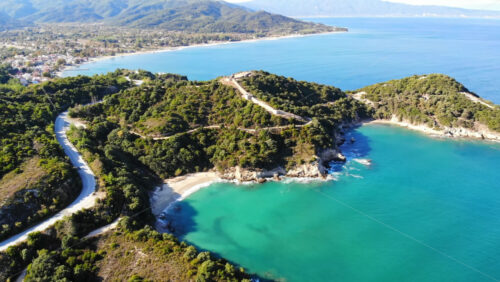 Video - Aerial drone view of the Aegean sea rocky coast, road and peninsula with greenery, ancient excavations on the hill, waves with a lot of foam, town on the background in Greece