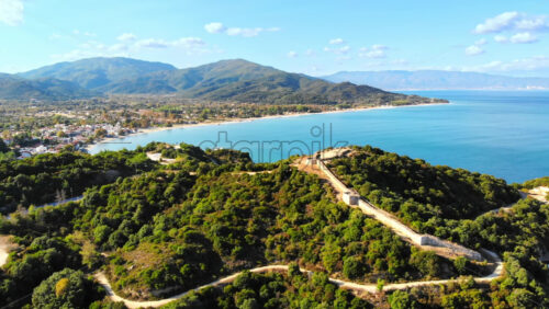 Video - Aerial drone view of the Aegean sea rocky coast, ancient wall excavations on a hill, town on the background in Greece