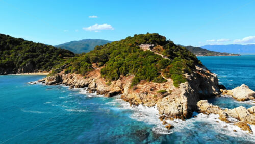 Video - Aerial drone view of the Aegean sea rocky coast, peninsula with greenery and ancient excavations on the hill, waves with a lot of foam, Greece