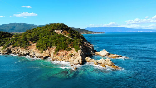 Video - Aerial drone view of the Aegean sea rocky coast, peninsula with greenery and ancient excavations on the hill, waves with a lot of foam, Greece