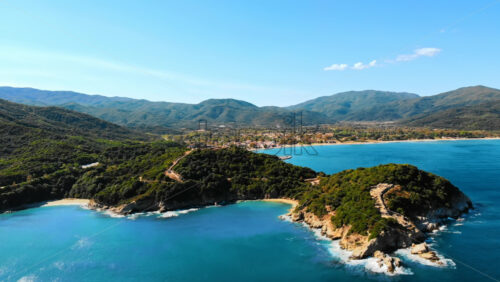 Video - Aerial drone view of the Aegean sea rocky coast, peninsula with greenery and ancient excavations on the hill, waves with a lot of foam, town on the background in Greece