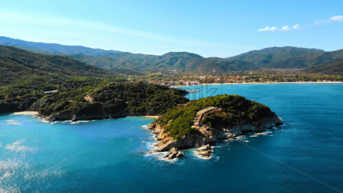 Video - Aerial drone view of the Aegean sea rocky coast, peninsula with greenery and ancient excavations on the hill, waves with a lot of foam, town on the background in Greece