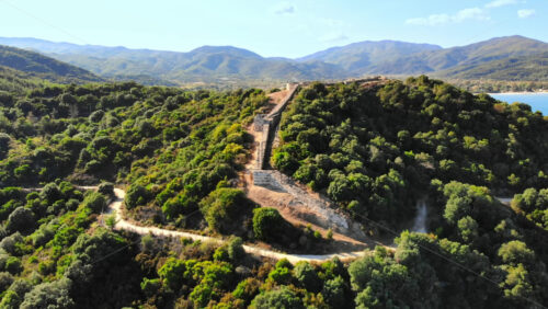 Video - Aerial drone view of the Aegean sea rocky coast, ancient wall excavations on a hill, town on the background in Greece