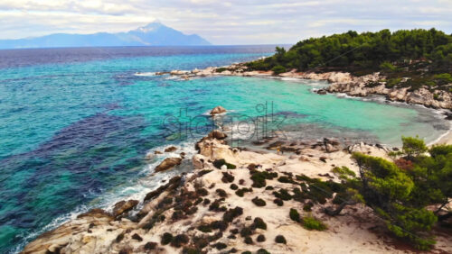 Video - Aegean sea rocky coast with greenery around, blue water and people on the beach, mountain in the distance, Greece