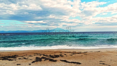 Video - Aerial drone view of Aegean sea coast in Greece. Waves and foam, blue water, land with a mountain on the background. Cloudy weather in Greece