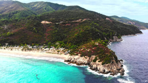 Video - Aerial drone view of the Aegean sea coast with camping near the beach, multiple greenery, hills on the background, blue water with waves. Greece