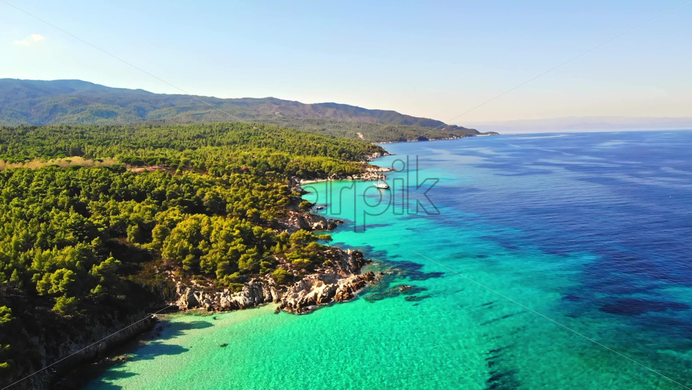 Video - Aerial drone view of the Aegean sea rocky coast with blue transparent water, multiple greenery, hills, moored sailboat. Greece