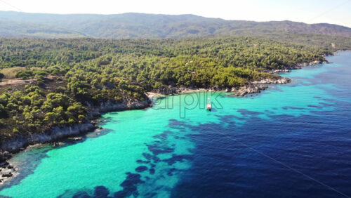 Video - Aerial drone view of the Aegean sea rocky coast with blue transparent water, multiple greenery, hills, moored sailboat. Greece