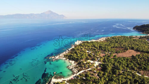 Video - Aerial drone view of the Aegean sea rocky coast with blue transparent water, multiple greenery, mountain on the background. Greece