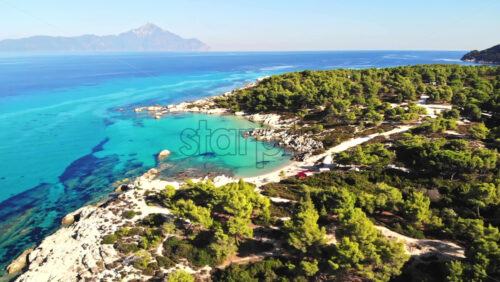 Video - Aerial drone view of the Aegean sea rocky coast with blue transparent water, multiple greenery, mountain on the background. Greece