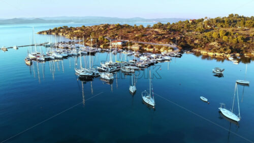 Video - Aerial drone view of Aegean sea port with multiple moored yachts at piers, greenery, blue water, sunset, Greece