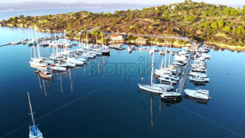 Video - Aerial drone view of Aegean sea port with multiple moored yachts at piers, greenery, blue water, sunset, Greece
