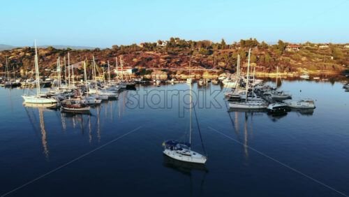 Video - Aerial drone view of Aegean sea port with multiple moored yachts at piers, greenery, blue water, sunset, Greece
