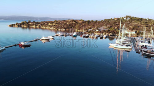 Video - Aerial drone view of Aegean sea port with multiple moored yachts at piers, greenery, blue water, sunset, Greece