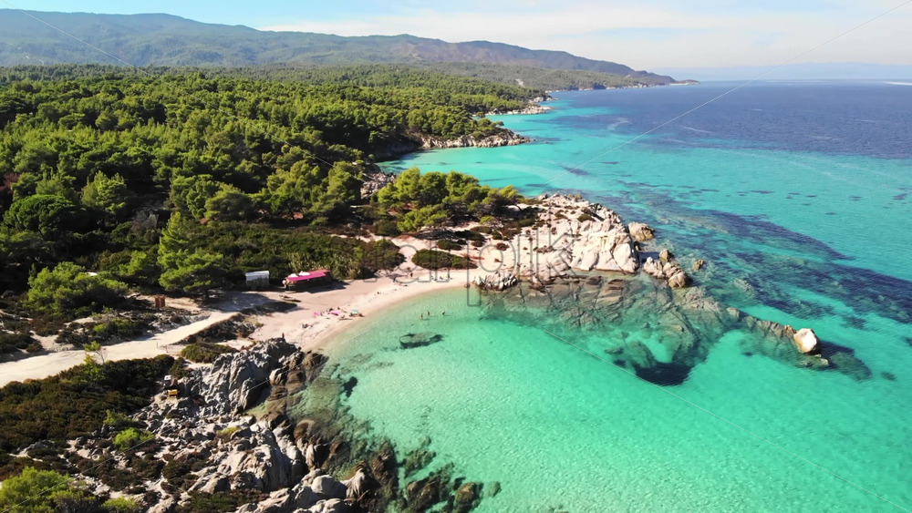 Video - Aerial drone view of the Aegean sea rocky coast with greenery around, blue water and resting people on the beach. Greece