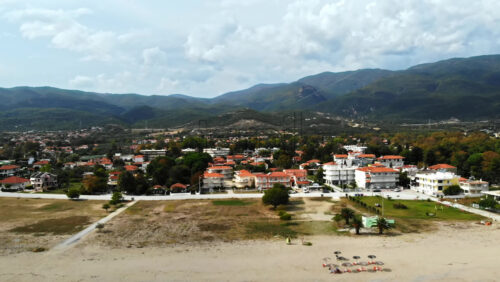 Video - Aerial drone view of the Aegean sea coast in Asprovalta, Greece. Beach with walking woman, umbrellas, multiple buildings and hills covered with greenery in the distance