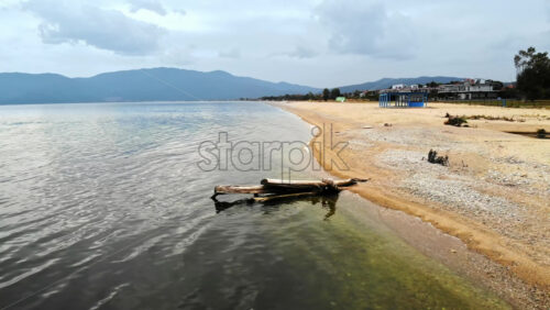 Video - View of the Aegean sea coast in Greece. Long beach with an old trunk in the water, small waves, buildings in the distance
