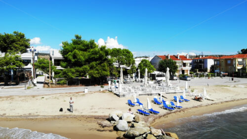 Video - Aerial drone view of the Aegean sea coast in Nikiti, Greece. A man contolling the drone, beach with teraces, umbrellas and sunbeds, multiple buildings