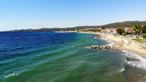 Video - Aerial drone view of the Aegean sea coast in Nikiti, Greece. Beach with waves of the sea, umbrellas, sunbeds greenery along the embankment street, multiple buildings