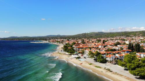 Video - Aerial drone view of the Aegean sea coast in Nikiti, Greece. Beach with waves of the sea, umbrellas, sunbeds greenery along the embankment street, multiple buildings, hills in th distance