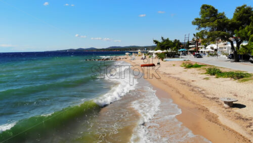 Video - Aerial drone view of the Aegean sea coast in Nikiti, Greece. Beach with waves of the sea, umbrellas, sunbeds greenery along the embankment street, multiple buildings