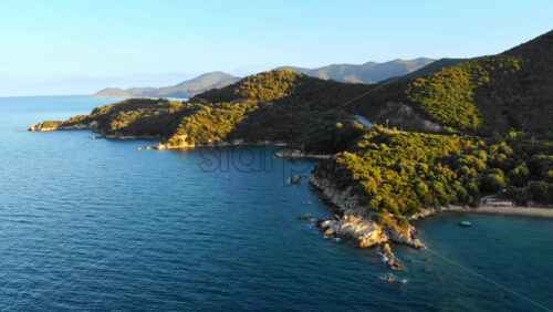 Video - Aerial drone view of the Aegean sea rocky coast with greenery and road on the shore, hills in the distance in Greece