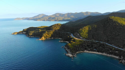 Video - Aerial drone view of the Aegean sea rocky coast with greenery and road on the shore, hills in the distance in Greece