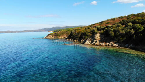 Video - Aerial drone view of the Aegean sea rocky coast with greenery along the shore and hills in the distance, blue water in Greece