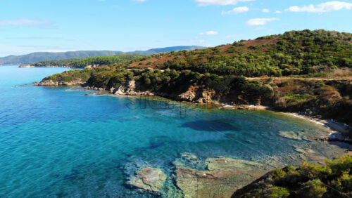 Video - Aerial drone view of the Aegean sea rocky coast with greenery along the shore and hills in the distance, blue water in Greece