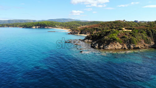 Video - Aerial drone view of the Aegean sea rocky coast with greenery and beaches along the shore and hills in the distance, blue water in Greece