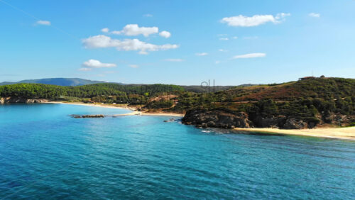 Video - Aerial drone view of the Aegean sea rocky coast with greenery and beaches along the shore and hills in the distance, blue water in Greece
