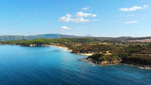 Video - Aerial drone view of the Aegean sea rocky coast with greenery and beaches along the shore and hills in the distance, blue water in Greece