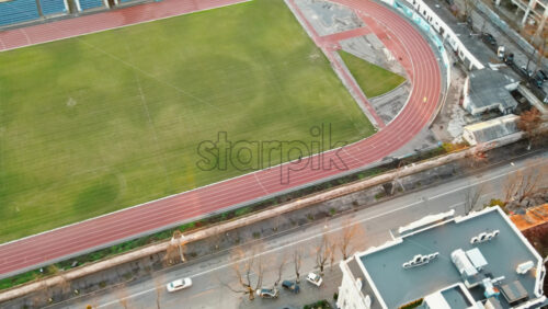 Video - Aerial drone view of a stadium in Chisinau. Green field, running tracks with a man, tribunes, buildings nearby, roads with moving cars and bare trees. Moldova