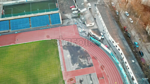 Video - Aerial drone view of a stadium in Chisinau. Green field, running tracks with a man, tribunes, buildings nearby, roads with moving cars and bare trees. Moldova