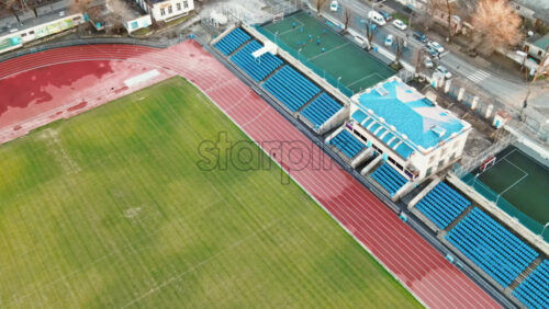 Video - Aerial drone view of a stadium in Chisinau. Green fields, running tracks with a man, tribunes, buildings nearby, roads with moving cars and bare trees. Moldova